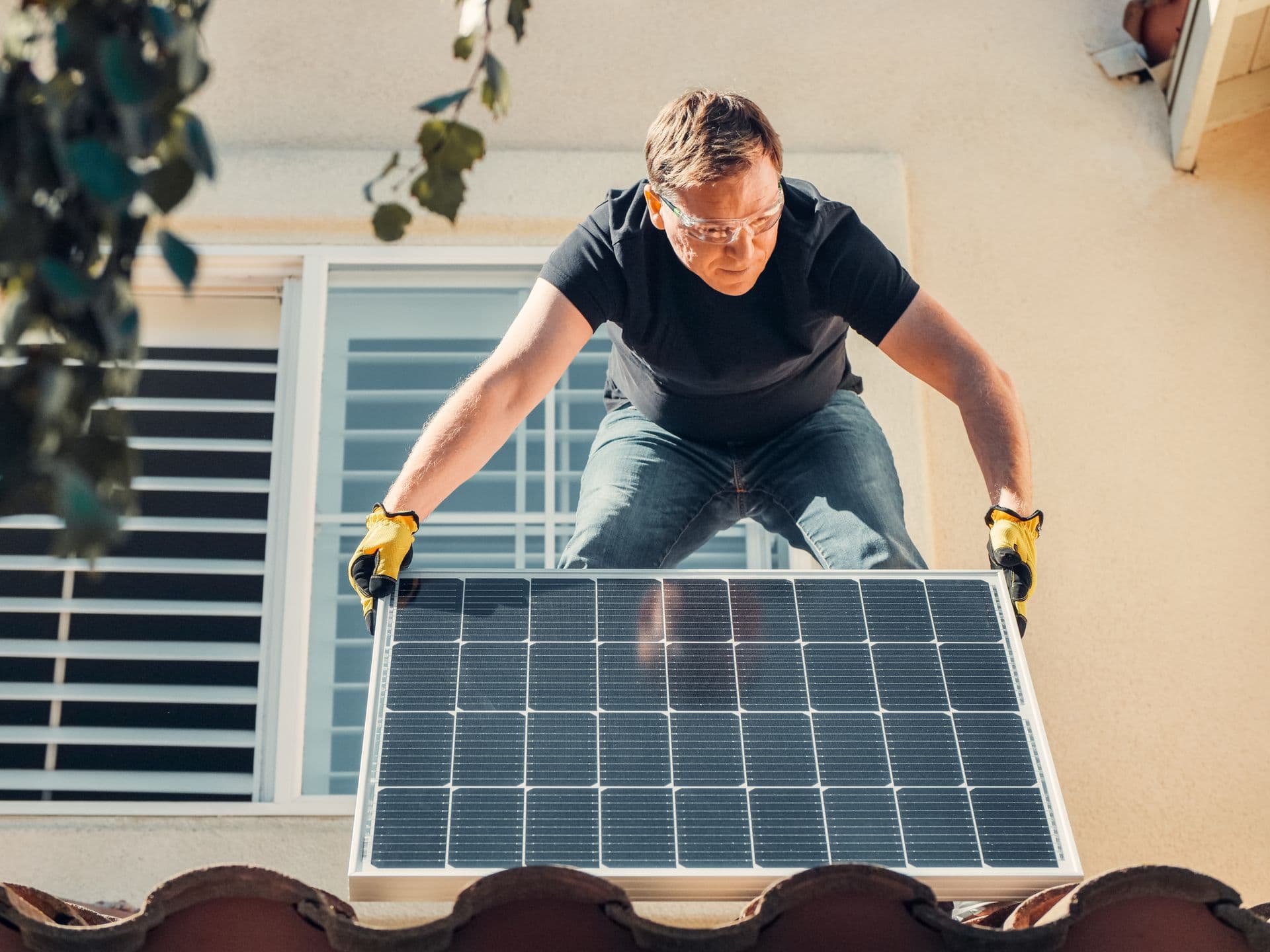A technician in safety glasses lowering a solar panel onto a tiled house roof